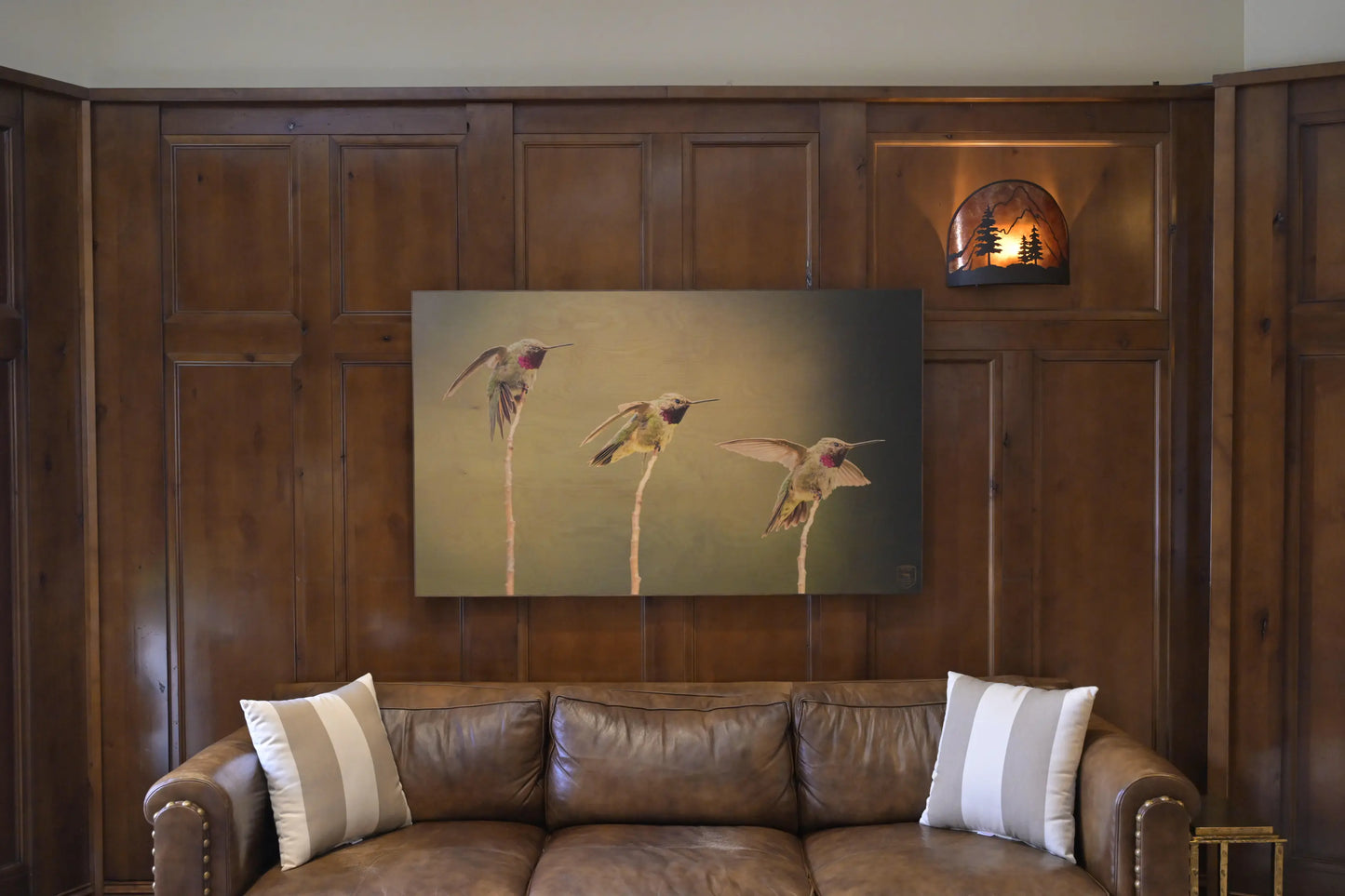 Brown leather sofa with decorative pillows in a room with wooden paneling and a painting of hummingbirds on the wall.
