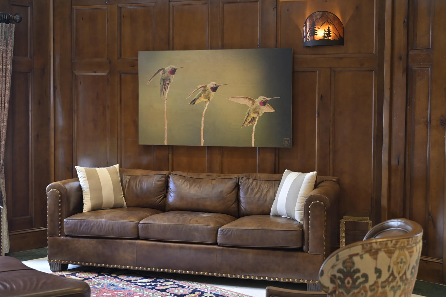 Brown leather sofa in a room with wooden walls and a painting of hummingbirds.