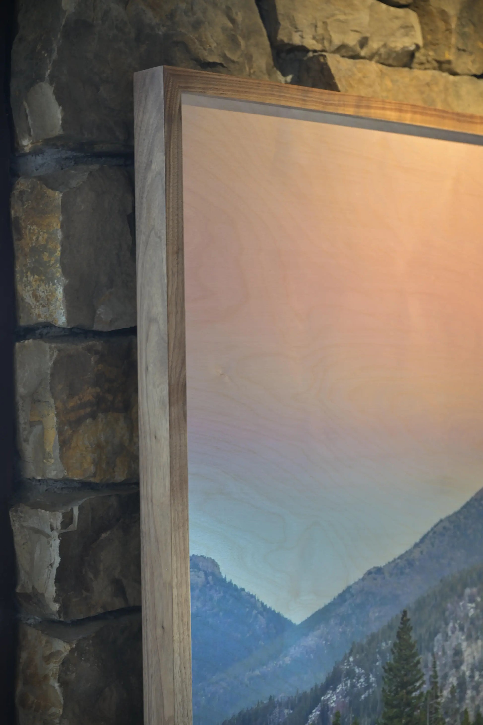 Wooden frame with a scenic mountain view inside, against a stone wall.