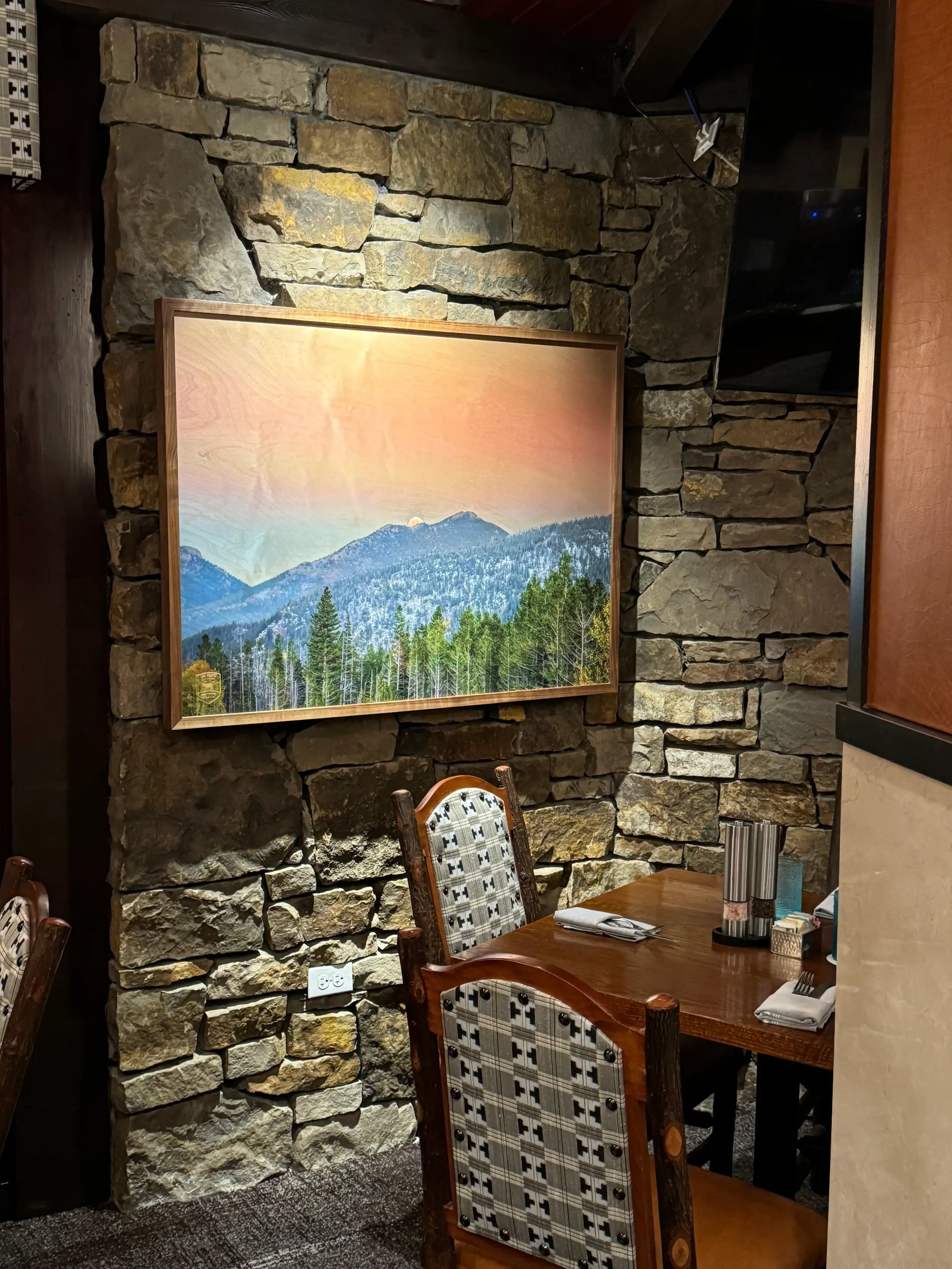 Framed landscape print on a stone wall above a wooden table with chairs.
