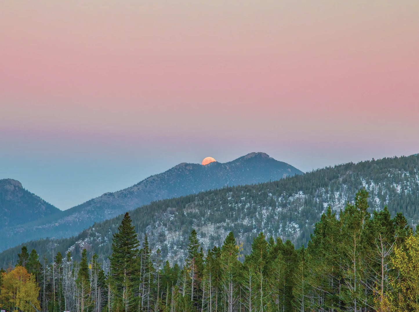 Mountain range with a pink and orange sunset in the sky, surrounded by trees.