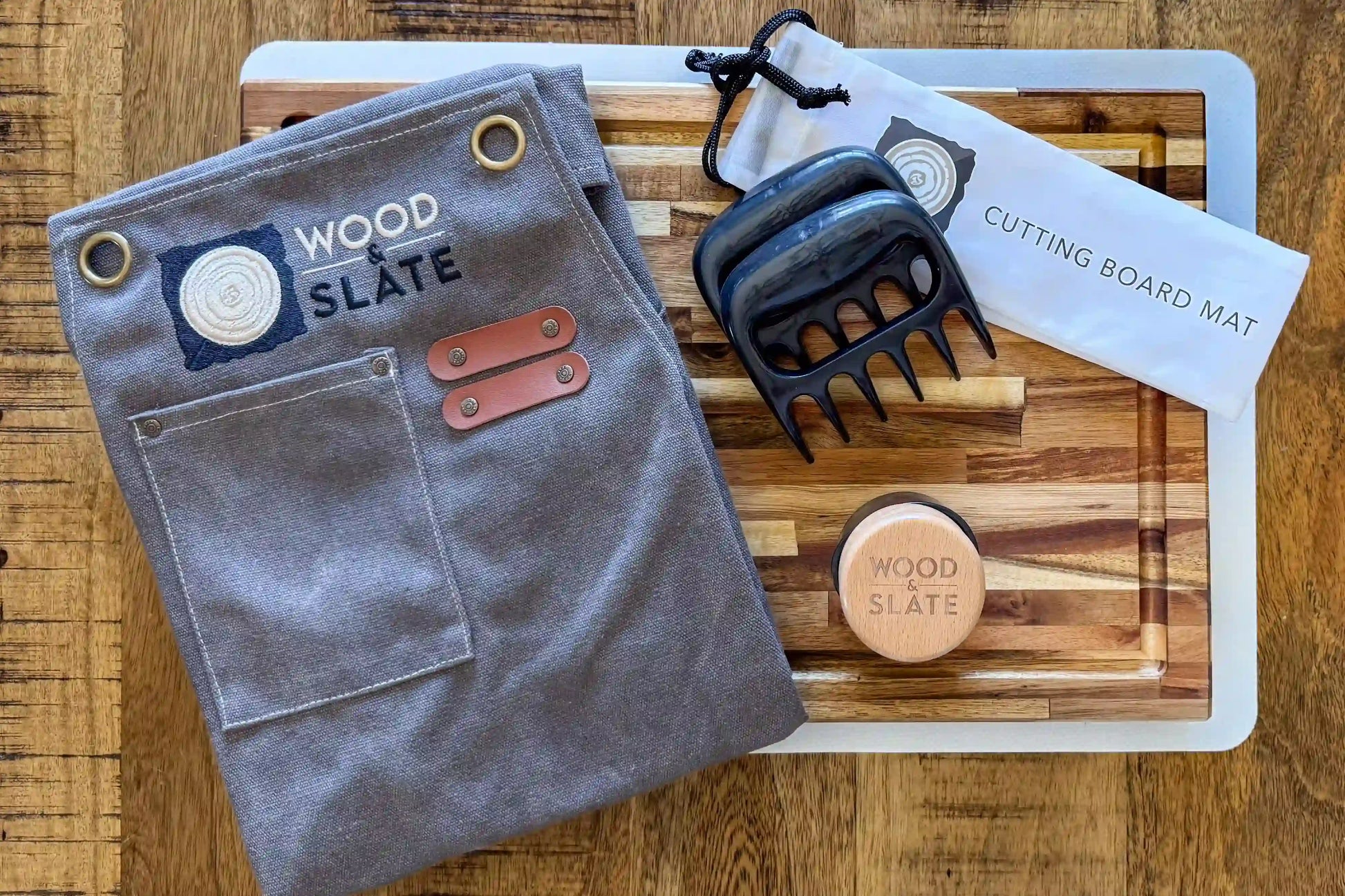Gray apron with leather accents, black meat tenderizer, and wooden soap on a cutting board mat.